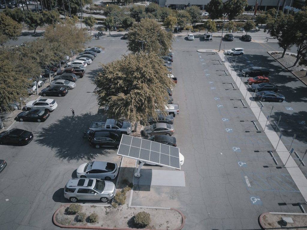 Aerial view of a solar panel charging station in a busy parking lot surrounded by cars and trees.